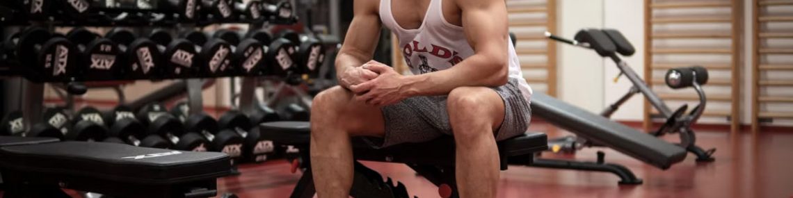 Strong, determined man in white tank top sitting in gym, showcasing fitness and strength.